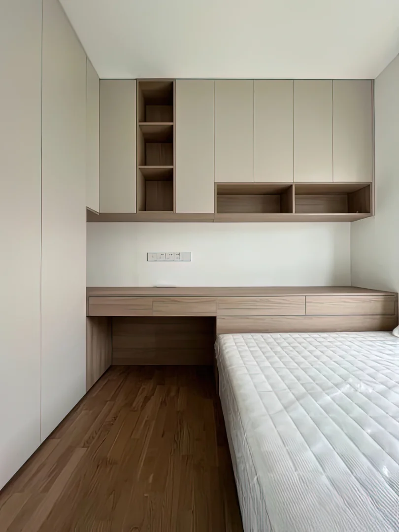 Compact HDB bedroom with a built-in wood study desk, bedside drawers and white overhead storage cabinets